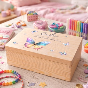 Può includere: Una scatola di legno con un disegno a farfalla e il nome "Emilia" stampato sul coperchio. La scatola è circondata da giocattoli per bambini, tra cui un unicorno di peluche, braccialetti e un diario. La scatola ha la data 24.10.2020 e misure.
