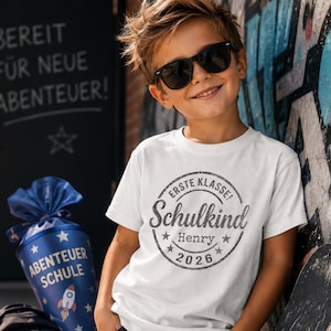 Könnte beinhalten: Ein weißes Kurzarm-T-Shirt mit einem kreisförmigen Aufdruck, der den Text "ERSTE KLASSE!", "Schulkind", "Henry" und "2026" umgeben von Sternen zeigt. Das T-Shirt wird mit einer dunklen Jeans, einer Sonnenbrille, einer Schultüte und einem Rucksack getragen.