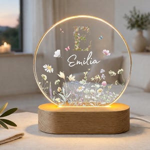 Könnte beinhalten: Ein kreisförmiges Acryl-Nachtlicht mit warmem LED-Schein, verziert mit einem Blumenmuster, dem Buchstaben E und dem in Weiß gedruckten Namen Emilia. Die Leuchte ist auf einem glatten, ovalen Holzsockel montiert, von dem ein weißes Stromkabel abgeht.