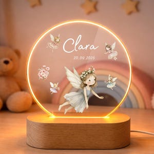 Peut inclure: Veilleuse personnalisée avec un design circulaire, une illustration de fée et le nom "Clara" et la date "20.09.2026". Elle possède une base en bois et un bord lumineux et chaleureux, créant une ambiance douce.