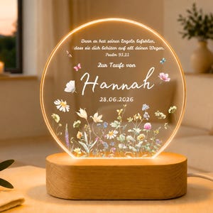 Könnte beinhalten: Ein kreisförmiges Acryl-Nachtlicht mit warm leuchtendem Rand auf einem hellen Holzsockel. Die Oberfläche zeigt einen bunten Wildblumen-Druck, Schmetterlinge und den Text: Denn er hat seinen Engeln befohlen, dass sie dich behüten auf all deinen Wegen. Psalm 91,11. Zur Taufe von Hannah 28.06.2026.