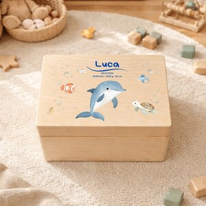 Könnte beinhalten: Helle Holz-Andenkenbox mit Delfin- und Meereskreaturen-Design. Die Box ist mit einem blauen Delfin, Fischen und einer Schildkröte verziert. Der Name "Luca" steht in blauer Schrift über dem Delfin, zusammen mit Datum, Uhrzeit, Gewicht und Größe.
