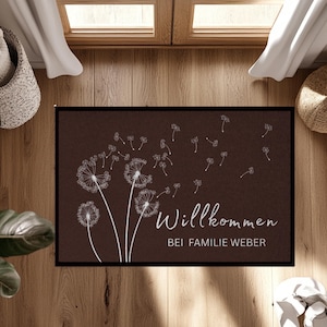 Könnte beinhalten: Eine rechteckige braune Fußmatte mit schwarzem Rand. Sie zeigt weiße Pusteblumen-Illustrationen und den deutschen Text "Willkommen BEI FAMILIE WEBER". Die Matte liegt auf einem Holzboden.