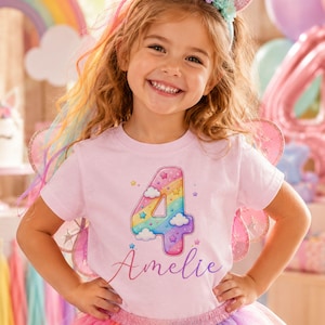 Puede incluir: Una camiseta de manga corta de color rosa claro con un gráfico grande del número 4 con degradado de arcoíris, nubes blancas y estrellas pequeñas. Debajo del número, el nombre Amelie está impreso en una fuente cursiva morada.