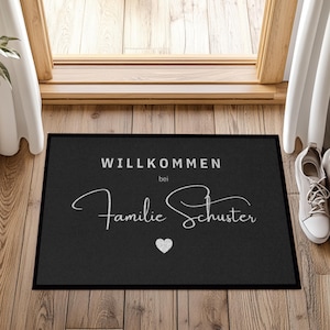 May include: Black rectangular doormat with white text that reads "WILLKOMMEN bei Familie Schuster" and a small heart. The mat is placed in front of a wooden door, with a pair of white sneakers to the right.