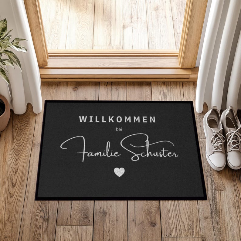 May include: Black rectangular doormat with white text that reads "WILLKOMMEN bei Familie Schuster" and a small heart. The mat is placed in front of a wooden door, with a pair of white sneakers to the right.