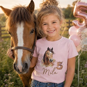 Può includere: Una t-shirt a maniche corte rosa chiaro con un'illustrazione in stile acquerello di un cavallo marrone, il numero 5 e il nome Mila stampato in corsivo marrone. La maglietta è indossata con un paio di jeans blu, creando un look casual.