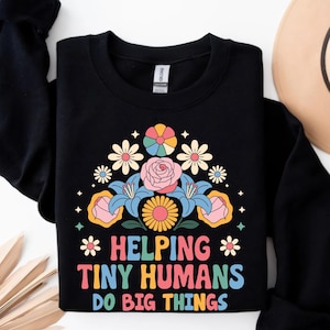 May include: Black sweatshirt with a floral design and the text "Helping Tiny Humans Do Big Things" in colorful letters. The floral arrangement includes pink roses, blue lilies, and daisies. A tan hat is visible in the background.