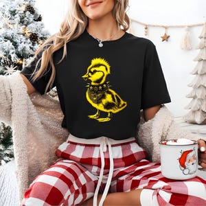 May include: Black t-shirt featuring a yellow punk rock duck graphic. The duck has a mohawk, a studded collar, and a piercing. The graphic is centered on the shirt. The shirt is paired with red and white plaid pajama pants.