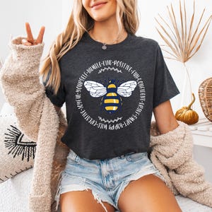 May include: Dark gray t-shirt featuring a stylized bee graphic with yellow and white wings and a yellow and black body. The text around the bee reads "WILD-CREATIVE-JOYFUL-YOURSELF-HUMBLE-KIND-POSITIVE-FUN-CONFIDENT-BRAVE-SMART-HAPPY".