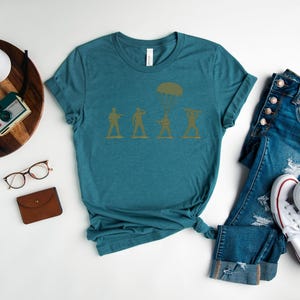 a casual outfit consisting of a teal t-shirt with a graphic design, a pair of blue jeans, white sneakers, and a pair of glasses. The outfit is laid out on a white surface, with a potted plant, a small camera, and a wallet nearby.