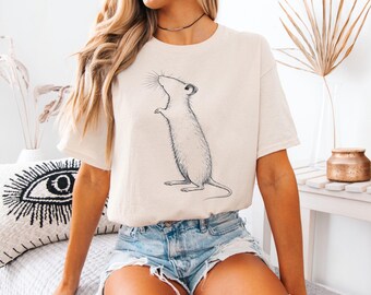 May include: Cream-colored t-shirt featuring a detailed black and white illustration of a rat standing upright. The rat is drawn in a realistic style, with visible whiskers and a long tail. The shirt has short sleeves and a relaxed fit.