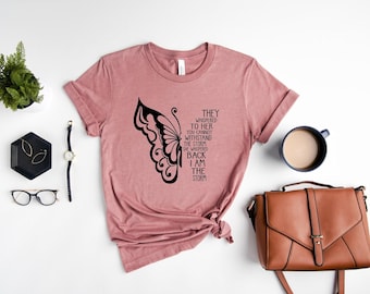 May include: A dusty rose t-shirt with a black butterfly wing design and the text "They whispered to her... I am the storm." Accessories include a watch, glasses, earrings, a purse, and a cup of coffee.