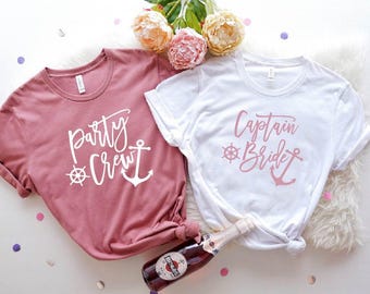 May include: Two t-shirts, one dusty rose and one white, with nautical-themed text. The dusty rose shirt says "Party Crew" with a ship's wheel and anchor. The white shirt says "Captain Bride" with a ship's wheel and anchor. A bottle of sparkling wine is in the image.