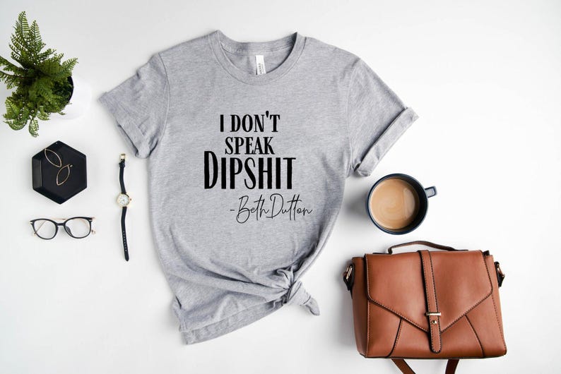 May include: A heather gray t-shirt with the text "I don't speak dipshit - Beth Dutton" in black. The shirt is styled with a brown purse, a watch, glasses, and a cup of coffee.