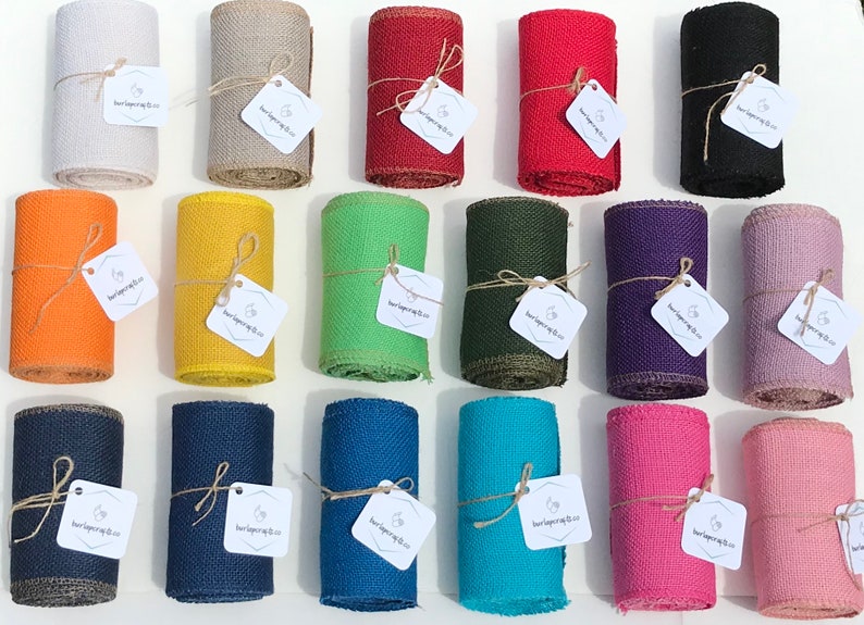 May include: A collection of rolled burlap ribbon in various colors, including white, tan, red, black, orange, yellow, green, dark green, purple, and pink. Each roll is tied with twine and has a tag attached.