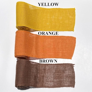 May include: Three rolls of burlap ribbon in yellow, orange, and brown. The text "YELLOW", "ORANGE", and "BROWN" are printed above each respective ribbon color. The ribbon is tightly rolled and appears to be made of a coarse, woven material.