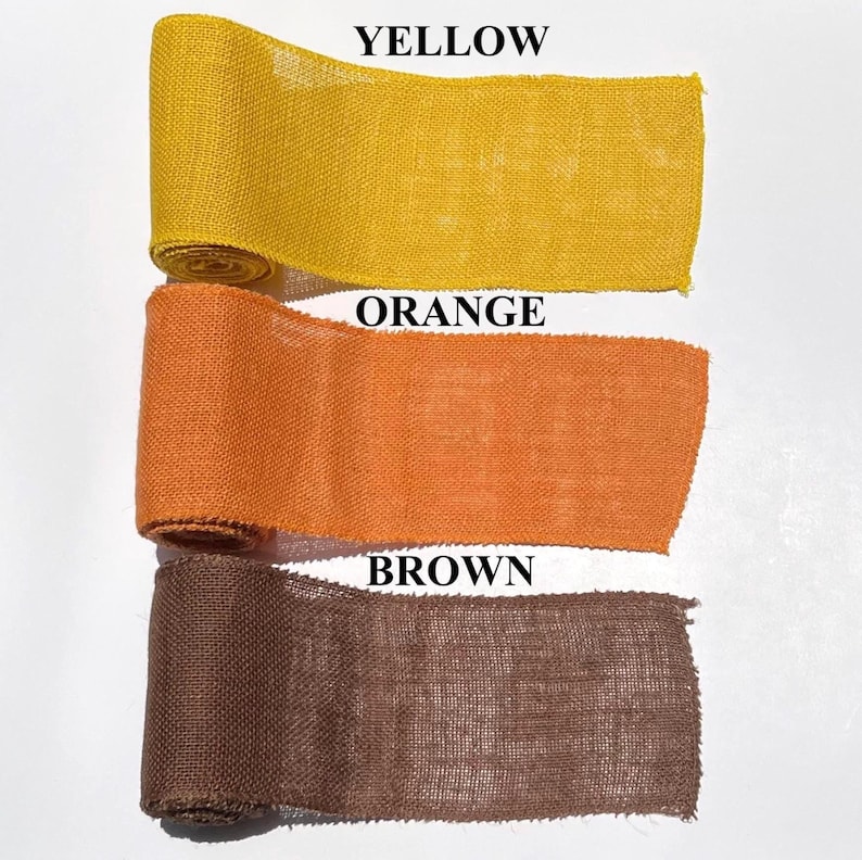 May include: Three rolls of burlap ribbon in yellow, orange, and brown. The text "YELLOW", "ORANGE", and "BROWN" are printed above each respective ribbon color. The ribbon is tightly rolled and appears to be made of a coarse, woven material.