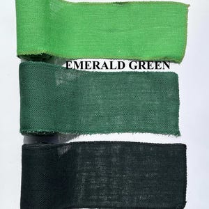 May include: Three rolls of burlap ribbon in shades of green and black. The top roll is apple green, the middle is emerald green, and the bottom is black. The text "APPLE GREEN", "EMERALD GREEN", and "BLACK" are printed above each ribbon.