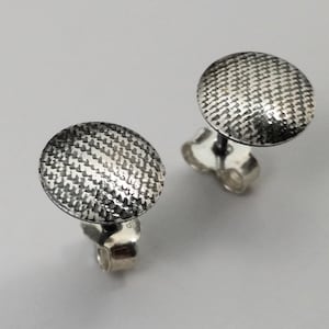 Può includere: Due orecchini a bottone in argento con una superficie circolare strutturata e un motivo intrecciato. Gli orecchini sono presentati su uno sfondo grigio chiaro, evidenziando la loro lucentezza metallica e il design dettagliato. Sono visibili anche i perni e i fermagli.