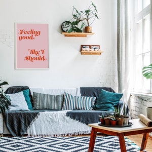 Puede incluir: Una impresi&oacute;n enmarcada con fondo rosa y marco blanco. La impresi&oacute;n presenta el texto "Feeling good, like I should." en letras rojas y naranjas. La impresi&oacute;n est&aacute; colgada en una pared de ladrillo blanco sobre un sof&aacute; gris con almohadas y una manta. Hay plantas y un estante de madera con art&iacute;culos decorativos en la pared.