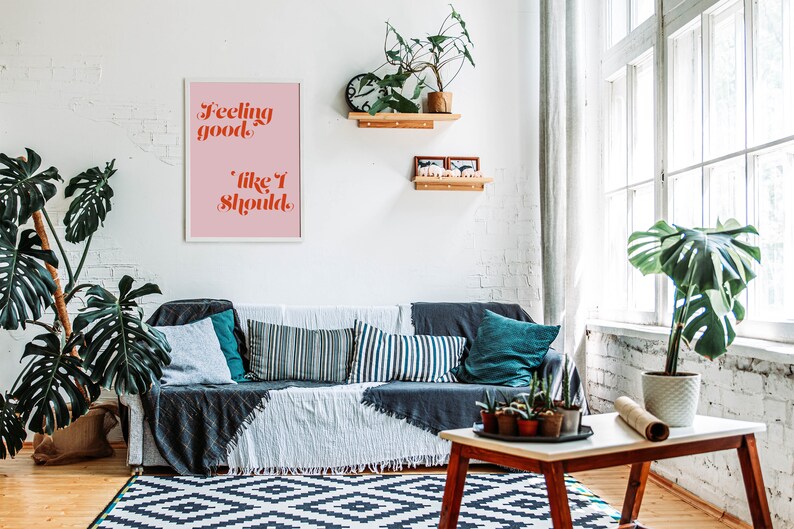 Puede incluir: Una impresi&oacute;n enmarcada con fondo rosa y marco blanco. La impresi&oacute;n presenta el texto "Feeling good, like I should." en letras rojas y naranjas. La impresi&oacute;n est&aacute; colgada en una pared de ladrillo blanco sobre un sof&aacute; gris con almohadas y una manta. Hay plantas y un estante de madera con art&iacute;culos decorativos en la pared.