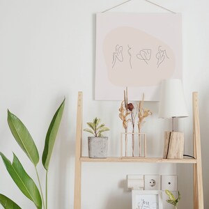 May include: A wooden shelf with two shelves, a white lamp with a white shade, a potted plant, a vase with dried flowers, a white mug, a framed print with the text "You belong among wildflowers", and a potted plant. The shelf is against a white wall with a framed print of a line drawing of three figures on a pink background.