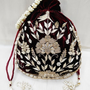 May include: A maroon velvet drawstring bag with gold and pearl embroidery featuring a floral design. The bag has a pearl chain handle and two pearl tassel accents.