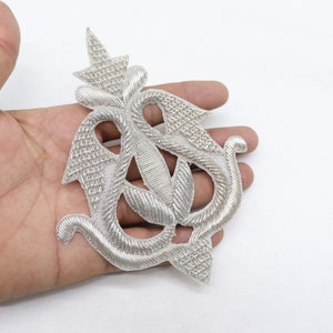 May include: A silver embroidered applique in the shape of a stylized floral design. The applique is intricate and detailed, with a raised texture.