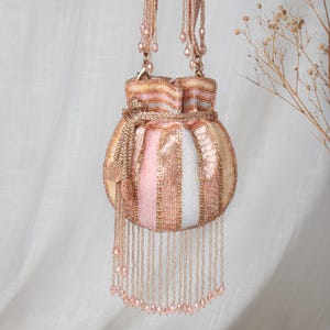 May include: A pastel-coloured beaded drawstring bag with pink, gold, and white stripes. It has a beaded handle and long beaded fringe. The bag is decorated with small, round, beaded embellishments.