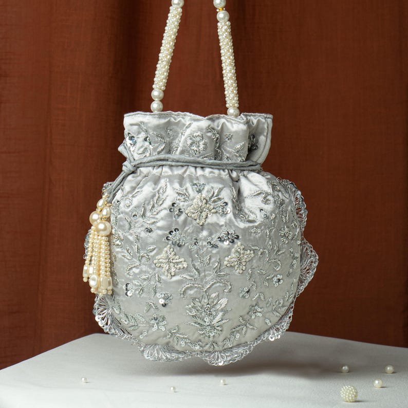 May include: A silver satin drawstring bag with intricate floral embroidery, sequins, and pearl accents. The bag features pearl beaded handles and a decorative pearl tassel. The bag is a formal accessory.