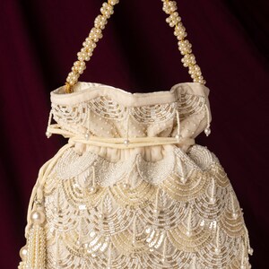 May include: A white beaded drawstring bag with a gold chain handle. The bag is decorated with pearl-like beads and sequins in a scalloped pattern.