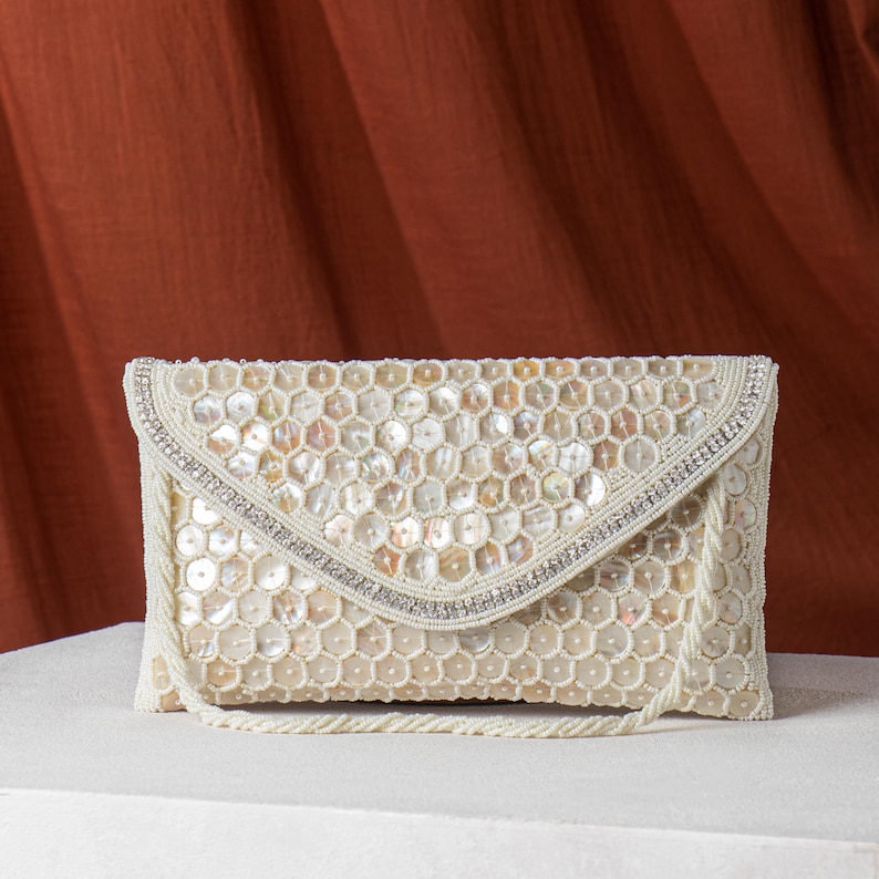 May include: Ivory-colored beaded clutch with a flap closure. The clutch is covered in iridescent, hexagonal sequins and features a beaded strap. A silver-toned band of rhinestones and beads accents the flap.