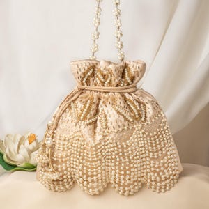 May include: A cream-coloured drawstring bag with a pearl and gold beaded design. The bag has a pearl chain strap and a scalloped bottom.