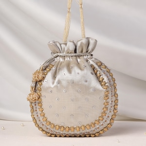 Puede incluir: Un bolso tipo saco de color plateado con cierre de cordón, parte superior fruncida y asas finas de cuerda. El exterior está decorado con pequeñas lentejuelas plateadas en cuadrícula y bordeado por un grueso adorno de flores metálicas doradas.