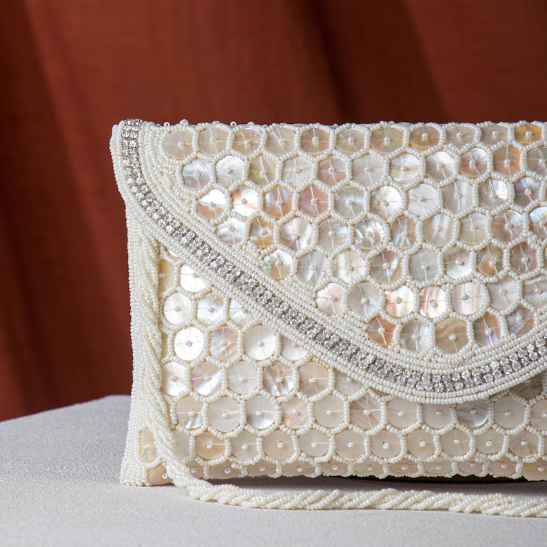 May include: Ivory-colored beaded clutch with a flap closure. The clutch features a honeycomb pattern of iridescent sequins and is trimmed with small white beads and a row of sparkling rhinestones. A matching beaded strap is attached.