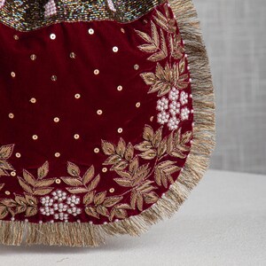 Könnte beinhalten: Nahaufnahme einer bordeauxroten Samt-Handtasche mit goldener Stickerei und Pailletten-Akzenten. Das Design zeigt goldene Blätter und weiße perlenartige Perlen. Der Rand ist mit goldenen Fransen verziert.