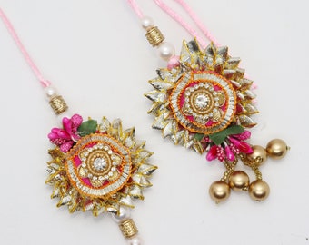 May include: Two pink and gold rakhi bracelets with decorative embellishments, including a flower design, beads, and a small bell. The bracelets are tied with pink thread.