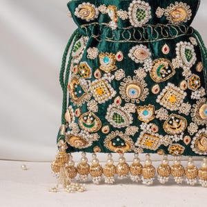 Könnte beinhalten: Ein dunkelgrüner Samtbeutel mit Kordelzug, verziert mit aufwendigen goldfarbenen Stickereien, kleinen weißen Perlen und bunten Steinen. Die Tasche ist mit einer unteren Fransenborte aus goldfarbenen Metallperlen und kleinen weißen Perlenclustern versehen.