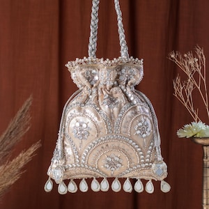 Peut inclure: Un sac beige à cordon de serrage orné de perles et de sequins argentés. Le sac est doté d'une poignée argentée tressée et d'un bord festonné avec des miroirs en forme de goutte d'eau. Le design est orné et élégant.