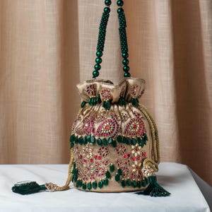 Könnte beinhalten: Ein goldfarbener Beutel mit Kordelzug und aufwendiger roter und goldener Stickerei. Die Tasche hat einen grünen Perlenhenkel und ist mit grünen Perlen und Quasten verziert. Das Design umfasst Blumen- und Paisley-Muster und schafft ein elegantes Accessoire.
