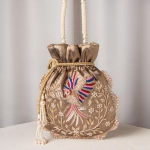 May include: A gold-colored drawstring bag with an embroidered bird design in pink, blue, and white. The bag has a pearl beaded handle and a tassel with pearl accents. The edges are scalloped with a lace trim.