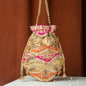 Könnte beinhalten: Eine verzierte Beuteltasche mit einem goldperlenbesetzten Henkel. Die Tasche weist aufwendige Stickereien in Rosa, Orange und Gold auf. Dekorative Elemente sind Blumen- und Vogelmotive mit goldenen Quasten am Boden. Die Tasche steht vor einem braunen Hintergrund.