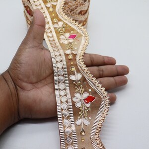 May include: A beige and gold embroidered trim with a scalloped edge. The trim features floral designs with red, pink, and white accents. The trim is embellished with sequins and beads.
