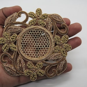 May include: A gold-colored, embroidered floral applique with a central circular mesh design. The applique features detailed leaves and flowers, creating an ornate, decorative piece. The design is held in a hand against a white background.