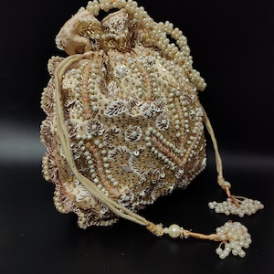 May include: A beige, beaded pouch with a drawstring closure. The pouch is adorned with white and gold beads, along with floral and leaf-like embellishments. A beaded handle and tassel accents complete the design. The bag is approximately 6 inches in diameter.