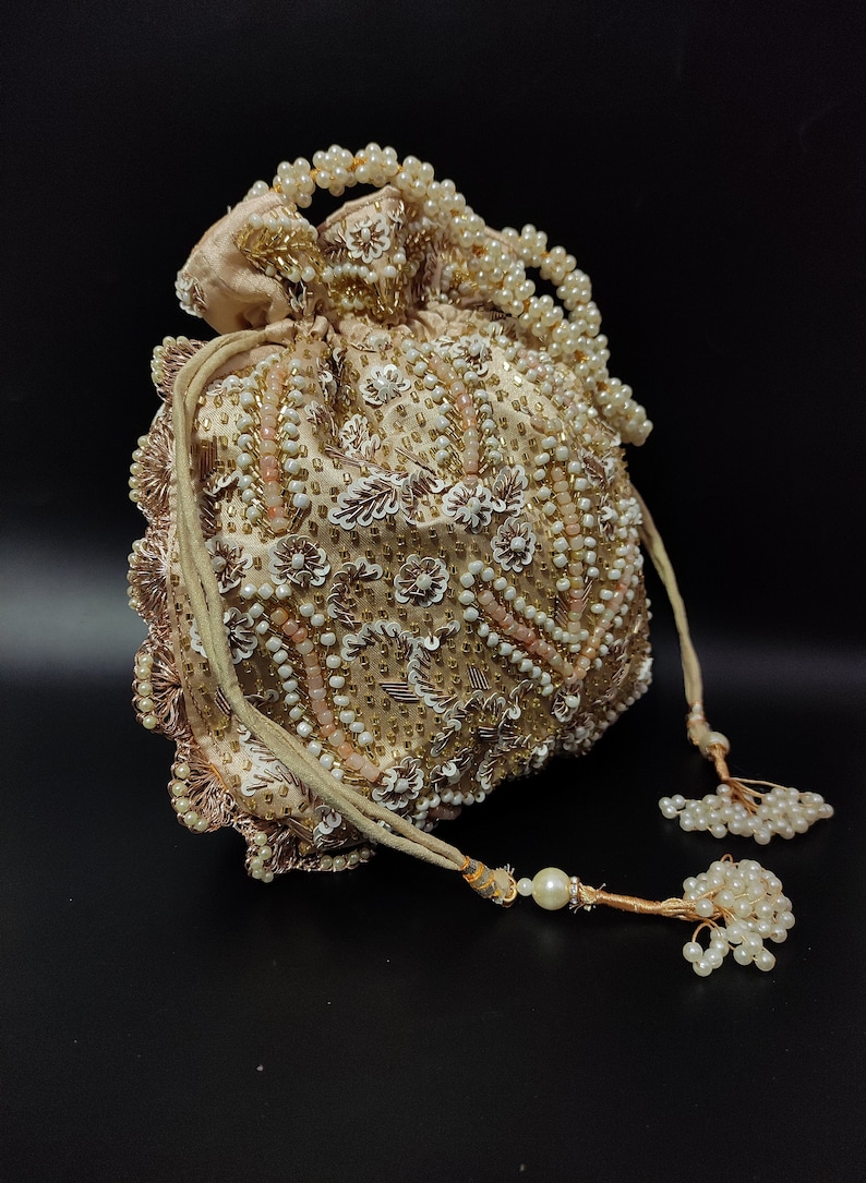 May include: A beige, beaded pouch with a drawstring closure. The pouch is adorned with white and gold beads, along with floral and leaf-like embellishments. A beaded handle and tassel accents complete the design. The bag is approximately 6 inches in diameter.
