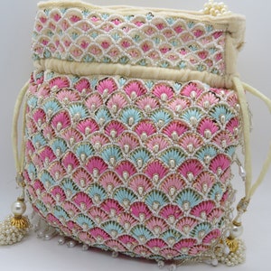 以下が含まれることがあります： ピンク、水色、白の刺繍が施されたクリーム色の巾着袋。 貝殻模様で、小さな真珠で装飾されています。 巾着から真珠のアクセントが付いたタッセルがぶら下がっています。