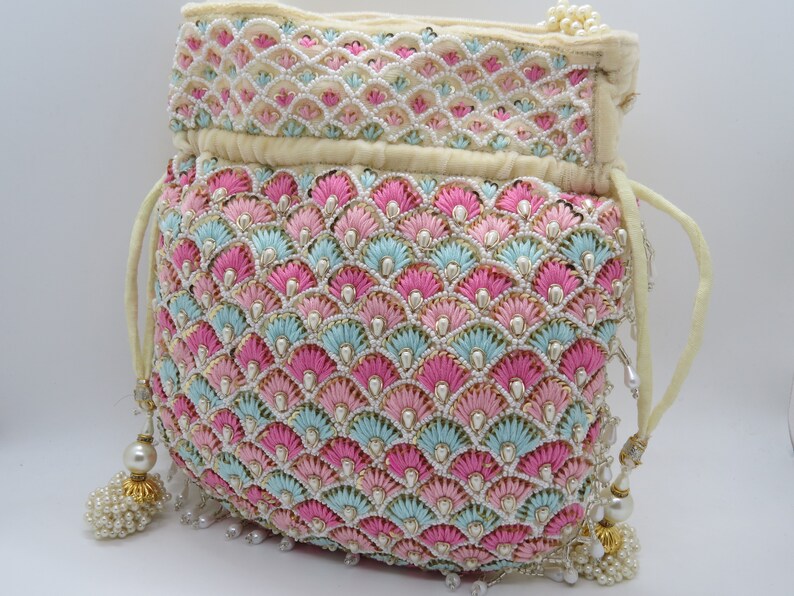 以下が含まれることがあります： ピンク、水色、白の刺繍が施されたクリーム色の巾着袋。 貝殻模様で、小さな真珠で装飾されています。 巾着から真珠のアクセントが付いたタッセルがぶら下がっています。