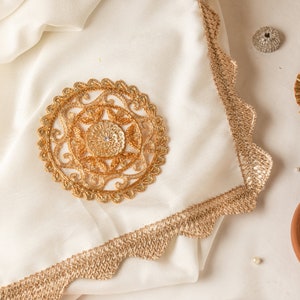 May include: A white fabric square with a gold embroidered design in the center. The fabric has a gold scalloped edge.